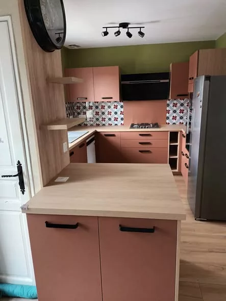 Vue d'ensemble de la cuisine présentant les façades terracotta rouge, les caissons en décor chêne naturel et les poignées noires. Le plan de travail entre l'évier et la plaque de cuisson, ainsi que le retour de l'îlot, offrent une surface particulièrement généreuse pour cuisiner.