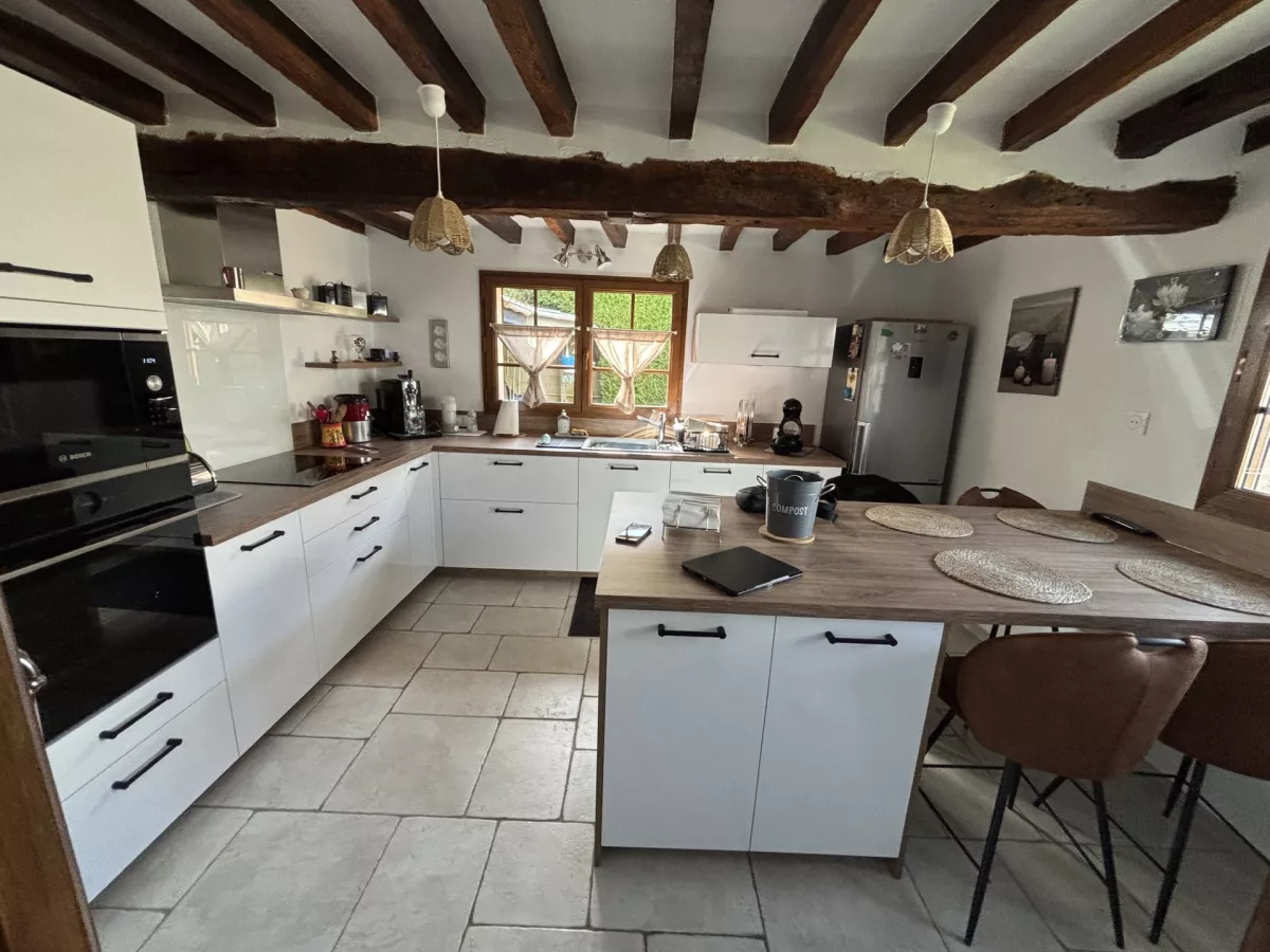 Vue d'ensemble de la cuisine aménagée avec un îlot central doté d'un plan de travail en décor chêne de Fontainebleau, des meubles bas à façades blanc royal brillant, un coin repas et des chaises hautes.