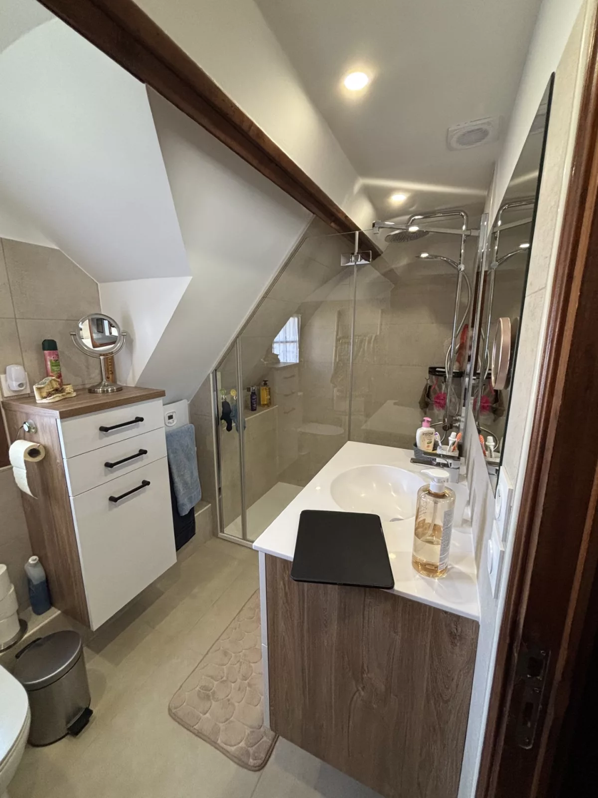 Autre angle de la pièce aménagée, montrant la douche avec sa paroi vitrée, le plan vasque et le meuble de rangement à tiroirs sous pente.